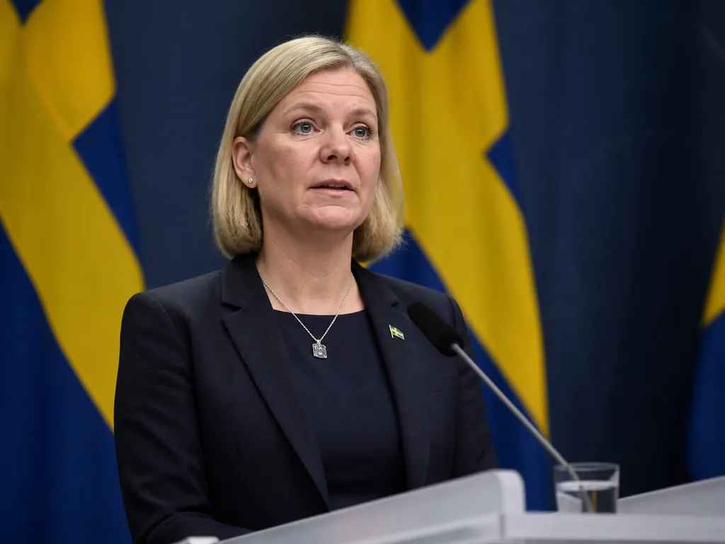 Statsminister vid talarpodium med svenska flaggor i bakgrunden